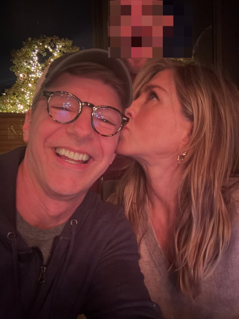 Jennifer Aniston in a grey sweater kissing Sean Hayes in a black hooded sweater and grey t-shirt on the cheek.