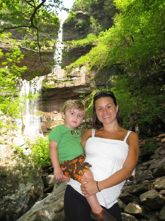 Rachel Napolitano-Curtis in a white tank top and black pants, holding Aidan Curtis, wearing a green t-shirt and brown and orange patterned shorts.