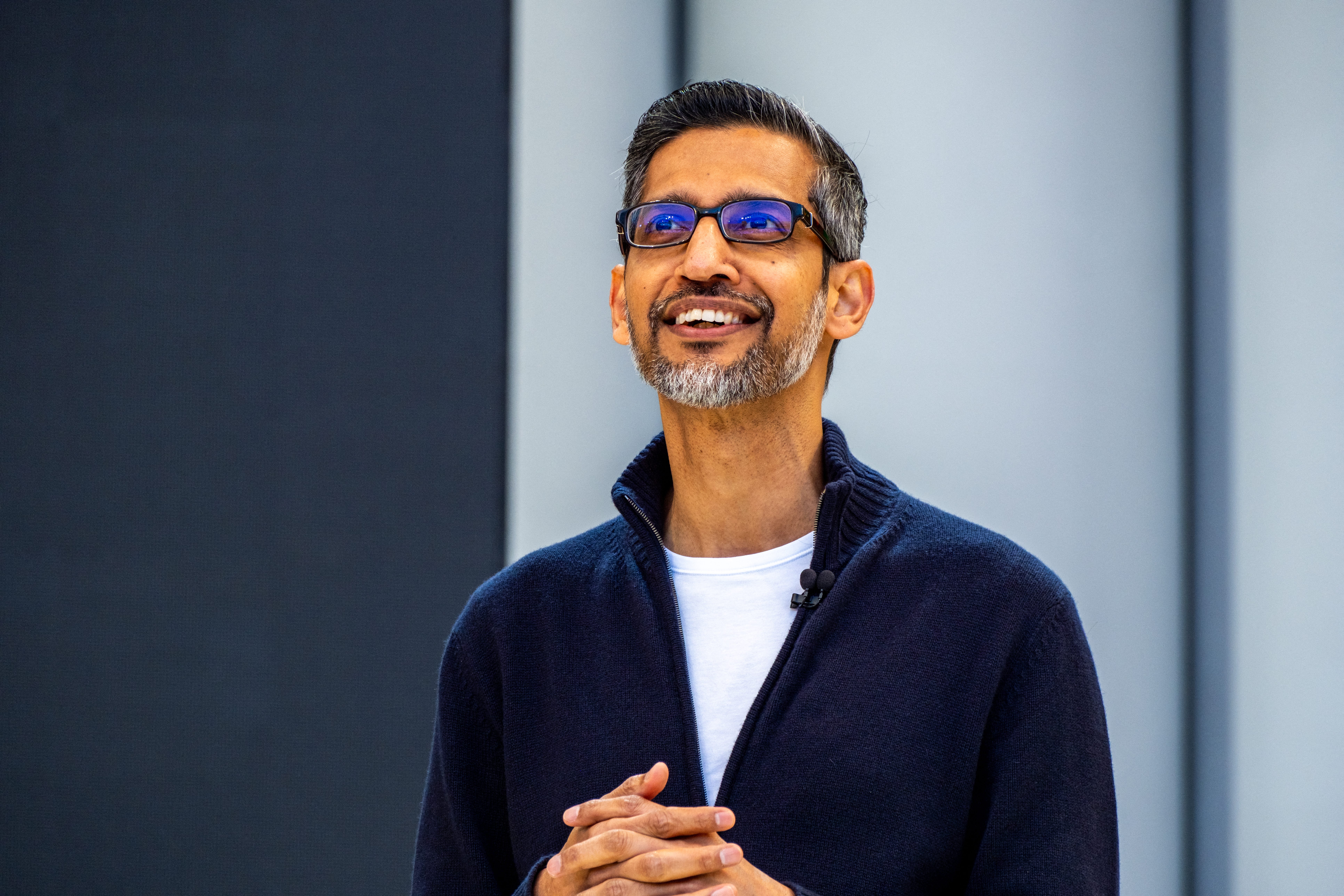 Google and Alphabet CEO Sundar Pichai looks upward, smiling, with his hands clasped in front of his stomach, wearing a blue zip sweater.