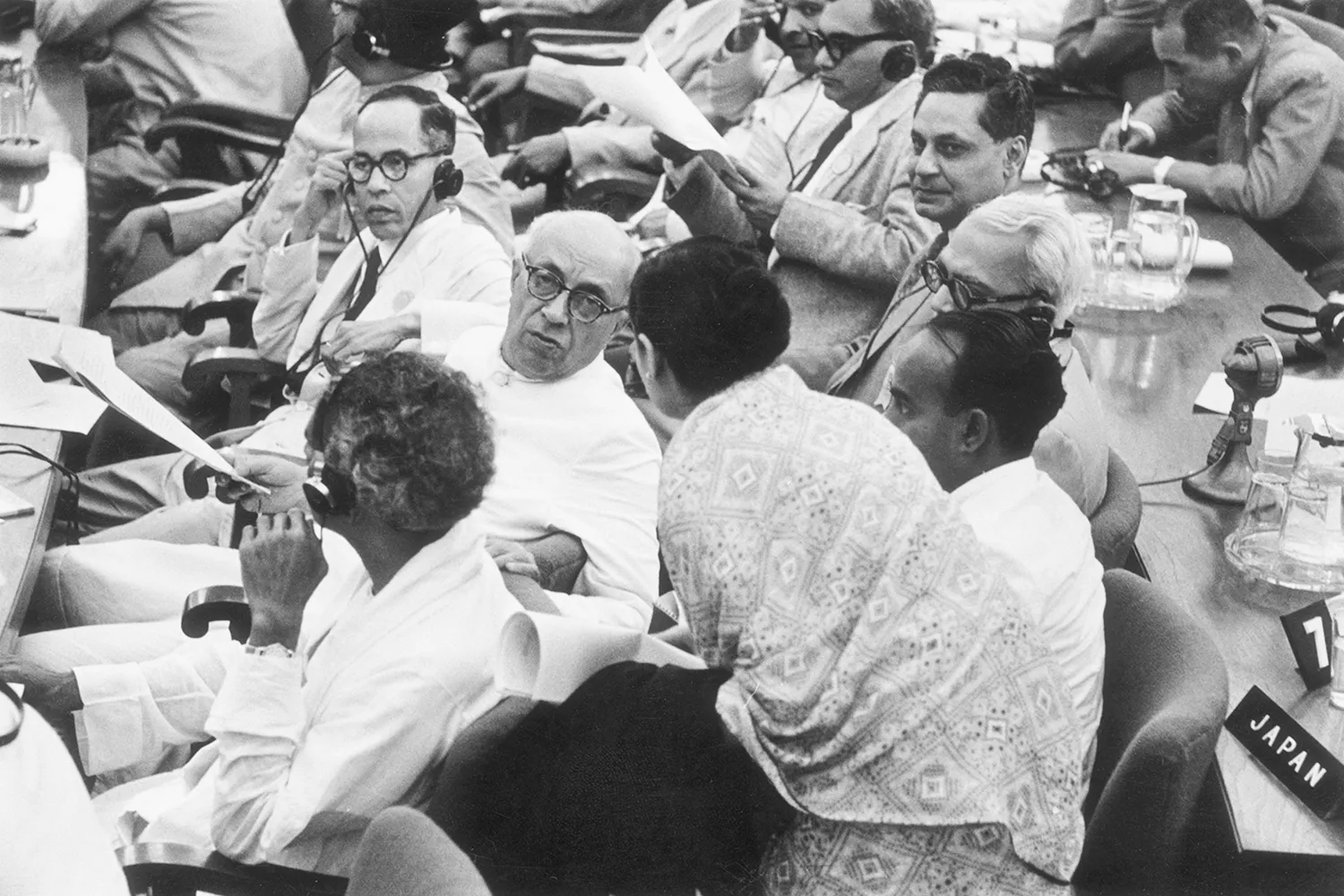 Nehru leands back to consult with another seated delegate in a crowd of people sitting in rows.