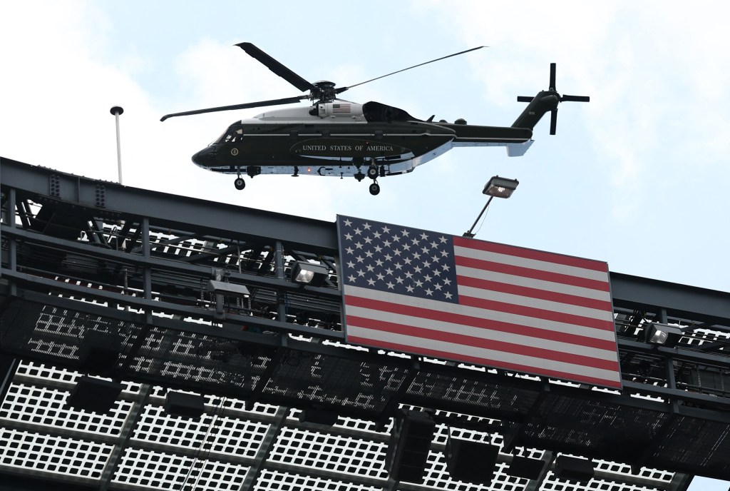Marine One helicopter over a stadium scoreboard displaying the American flag.