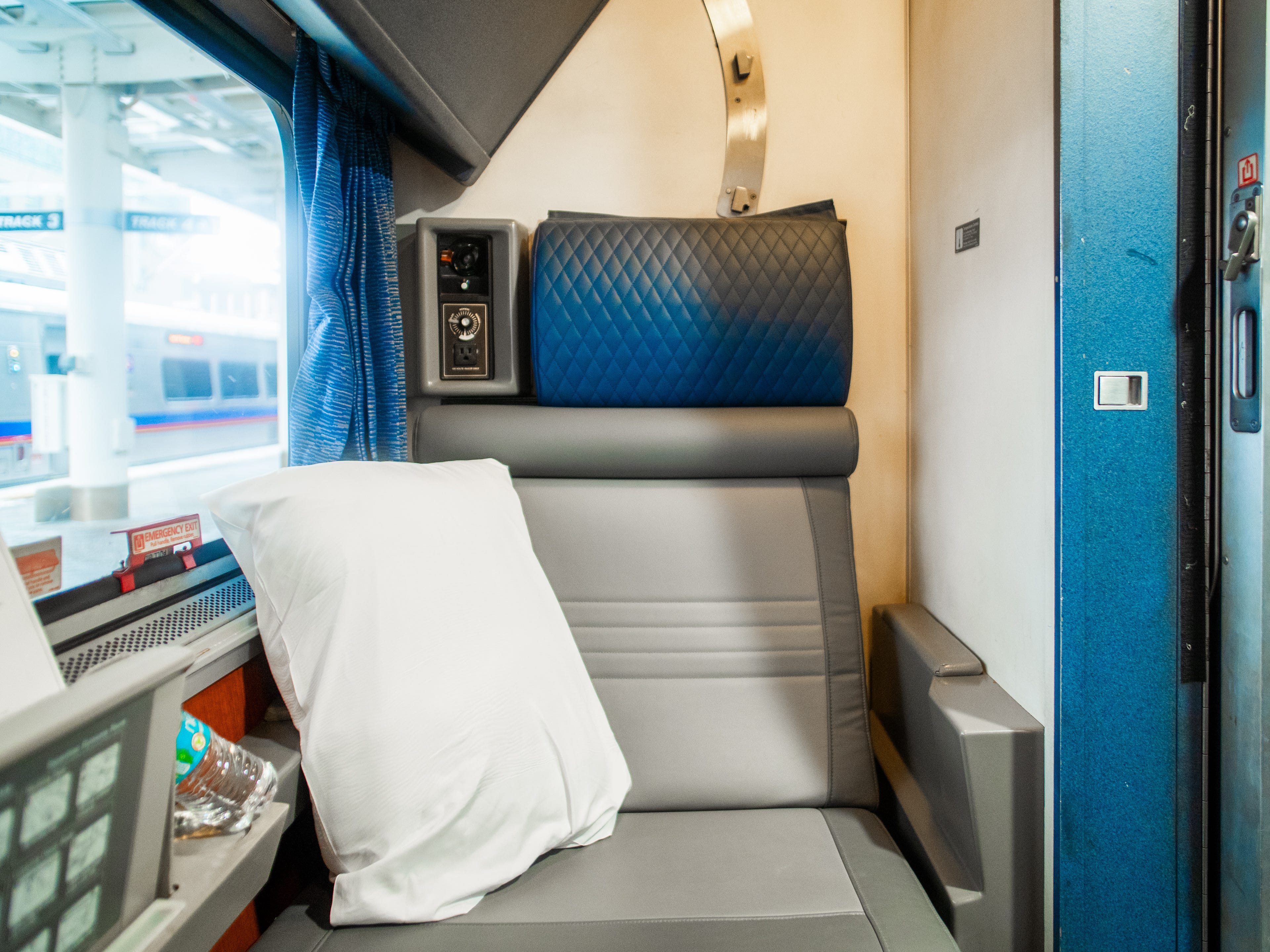 A white pillow sites on a gray train seat with a blue headrest inside an Amtrak roomette