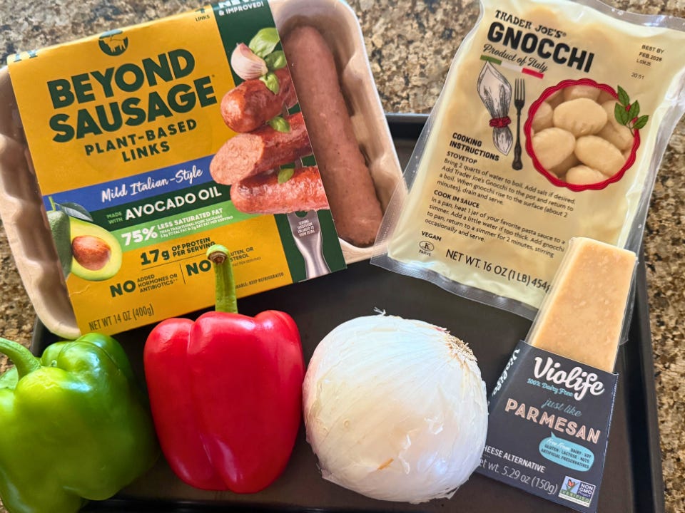 A package of vegan sausage, two bell peppers, an onion, a package of Trader Joe's gnocchi, and a block of Parmesan cheese on a countertop.