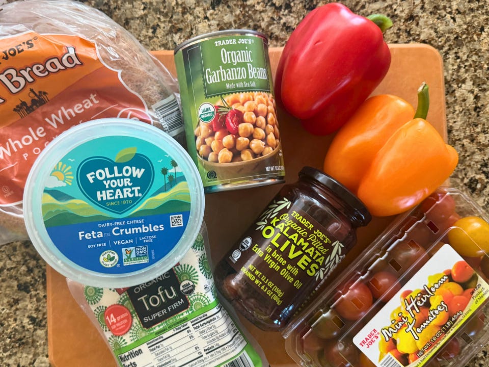 A package of Trader Joe's pita bread, a tub of feta crumbles, a package of tofu, a jar of garbanzo beans, a jar of kalamata olives, two bell peppers, and a container of heirloom tomatoes on a countertop.