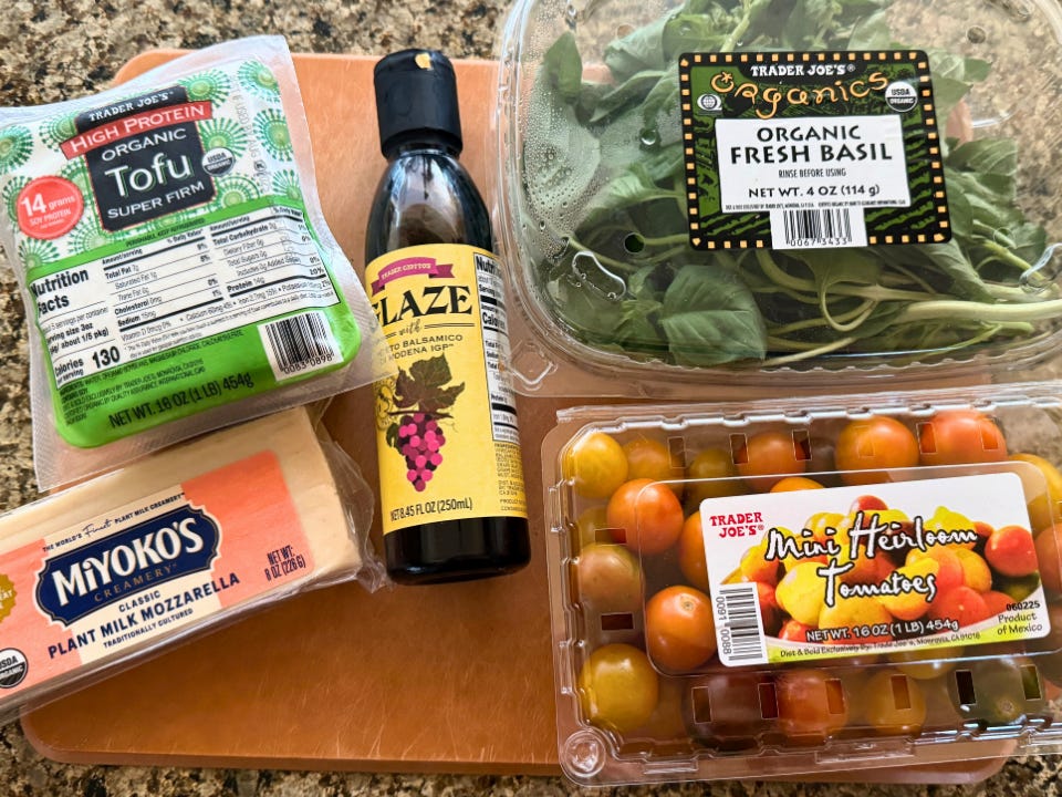 A package of tofu, a block of vegan mozzarella, a bottle of balsamic glaze, a container of fresh basil, and a container of heirloom tomatoes on a countertop.