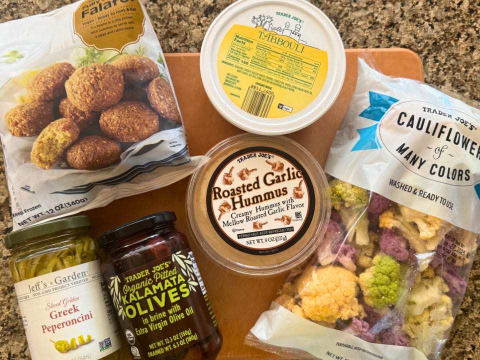 A bag of frozen falafel, a jar of pepperoncini, a jar of kalamata olives, a container of tabbouleh, a tub of garlic hummus, and a bag of cauliflower florets on a countertop.