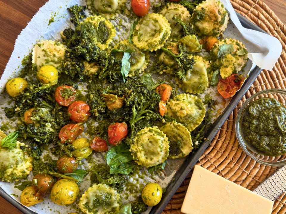 A sheet pan with Broccolini, tomatoes, ravioli, and pesto with a black of Parmesan on the side.