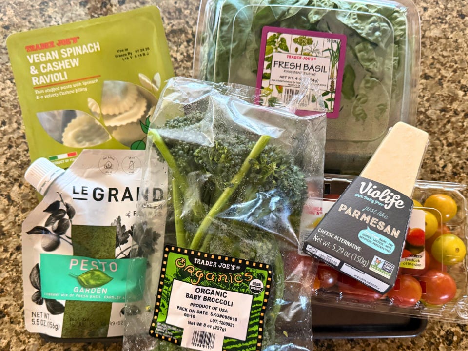 A package of vegan Trader Joe's ravioli, a pouch of pesto, a bag of Broccolini, a container of basil, a block of Parmesan, and a container of tomatoes on a countertop.