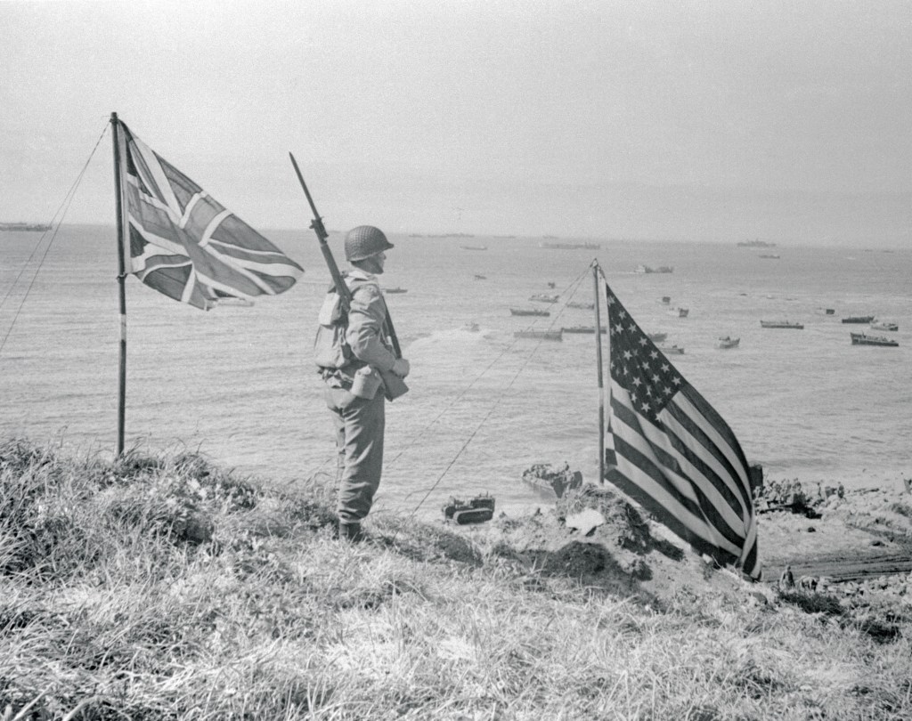 A soldier overlooking a beach landing, with the American and British flags displayed.