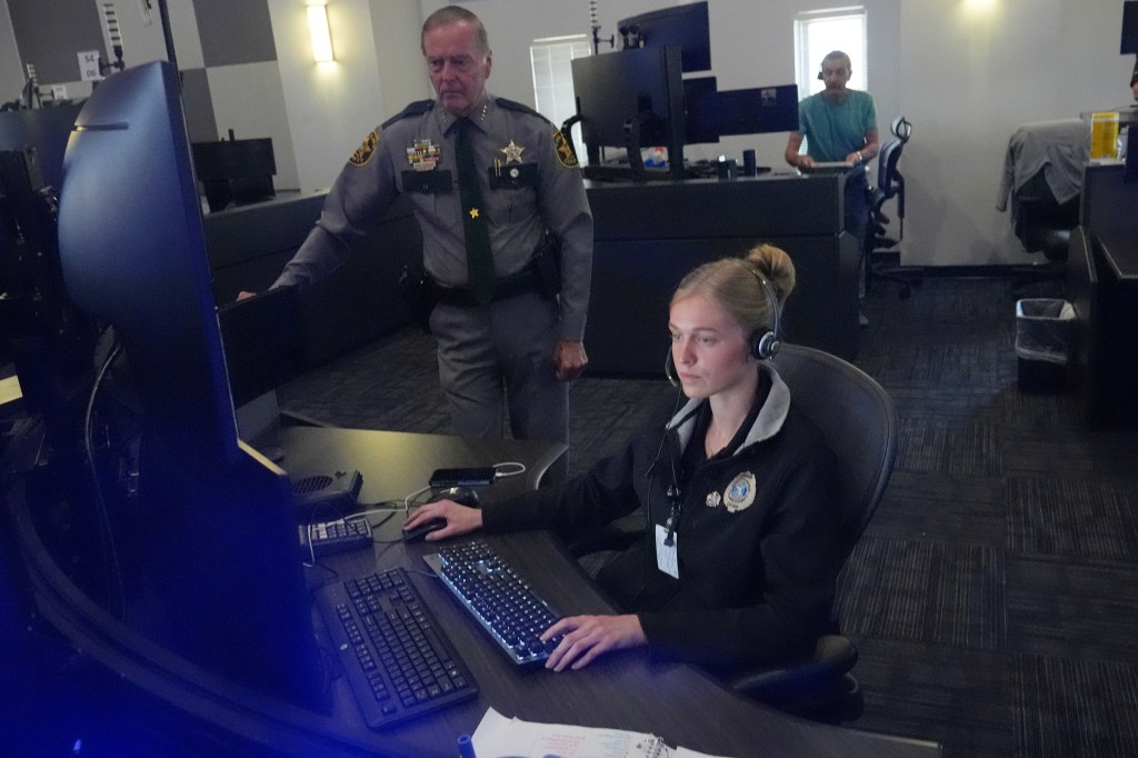 A female 911 dispatcher works at a computer console, with a sheriff standing nearby.