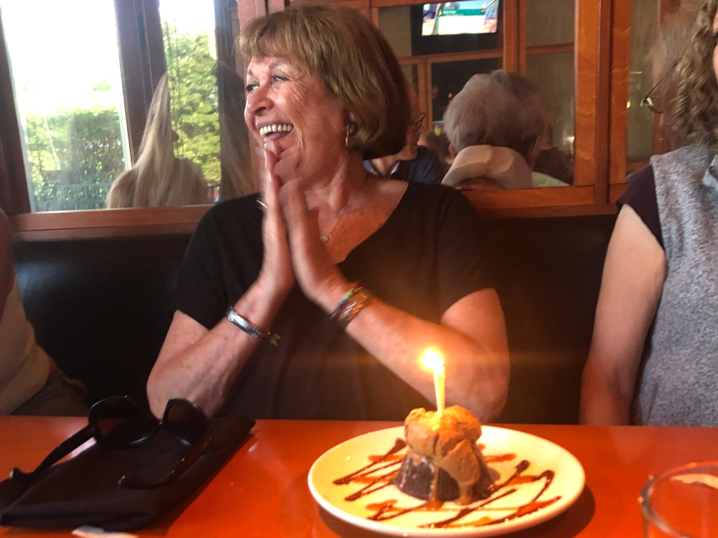 Author's grandmother clapping at a restaurant, smiling, with candle in dessert on table in front of her