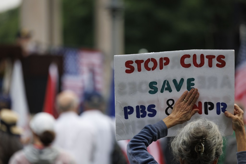 A demonstrator displays a sign that reads 'Stop Cuts Save PBS & NPR,' while taking part in a rally on the Boston Common on June 6, 2025.