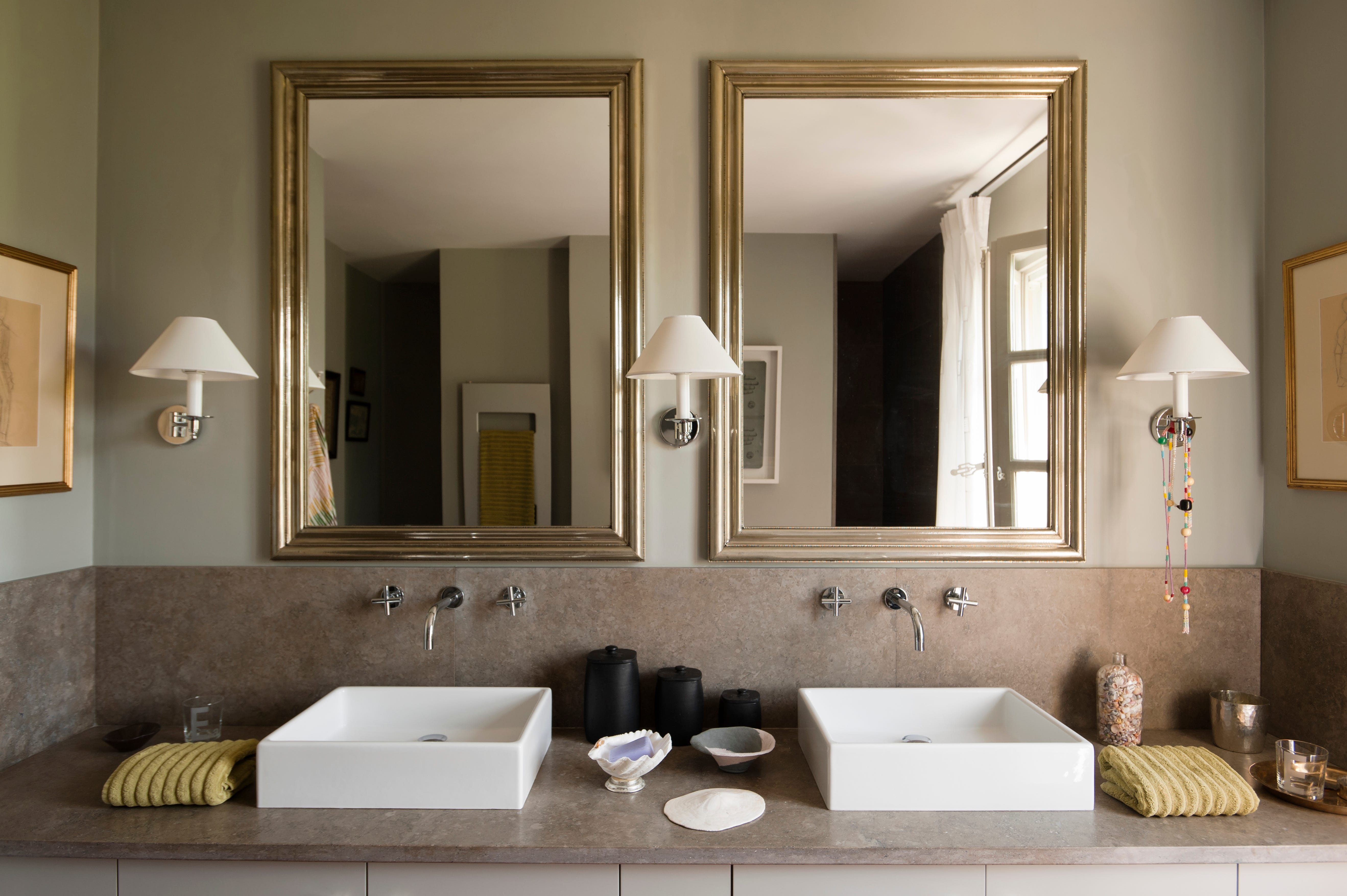 Sconces on a gray bathroom wall in between two mirrors with gold frames.