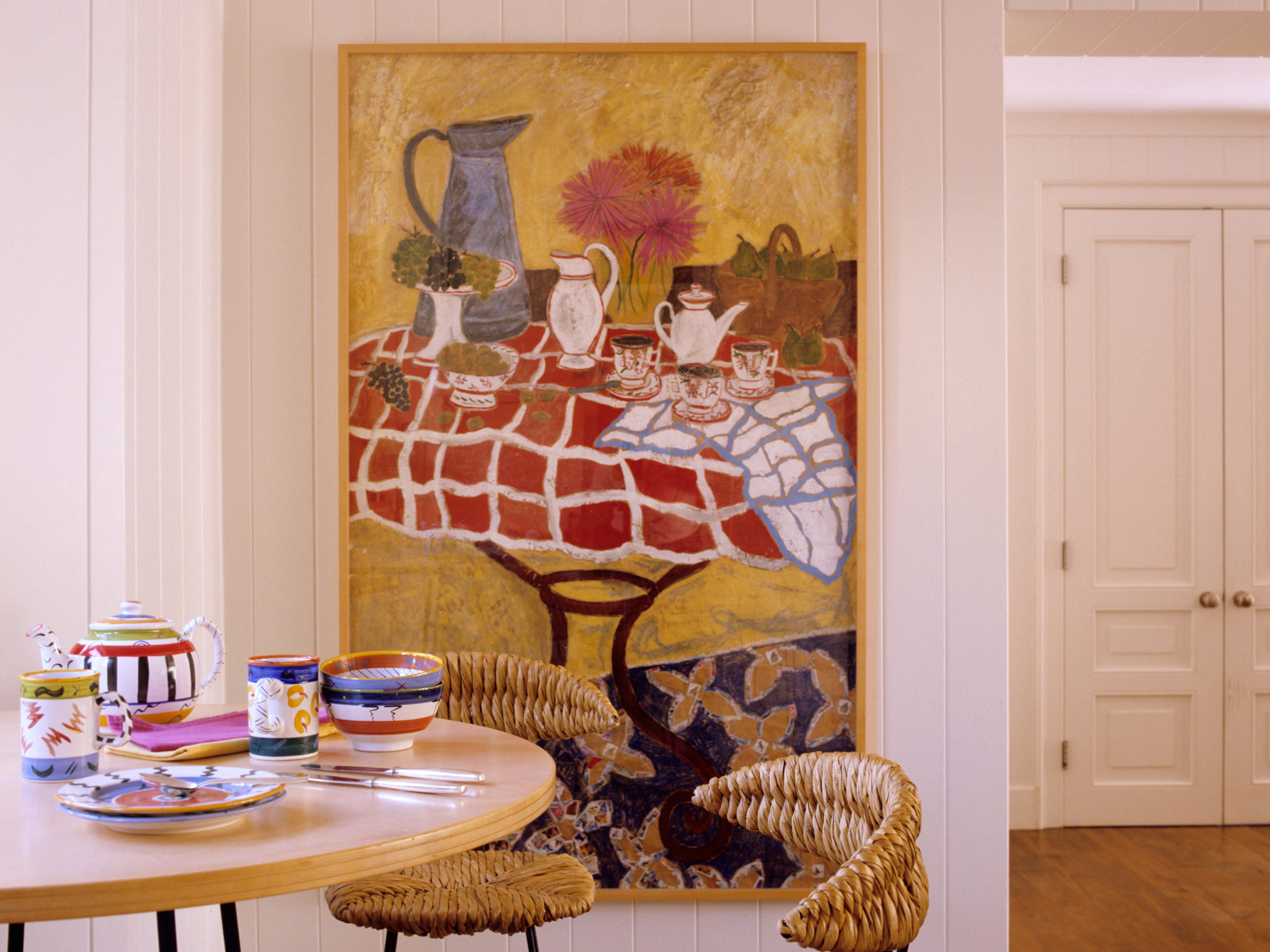 A room with a dining table with colorful dishes and a large painting of a dining table with teapots and flowers.