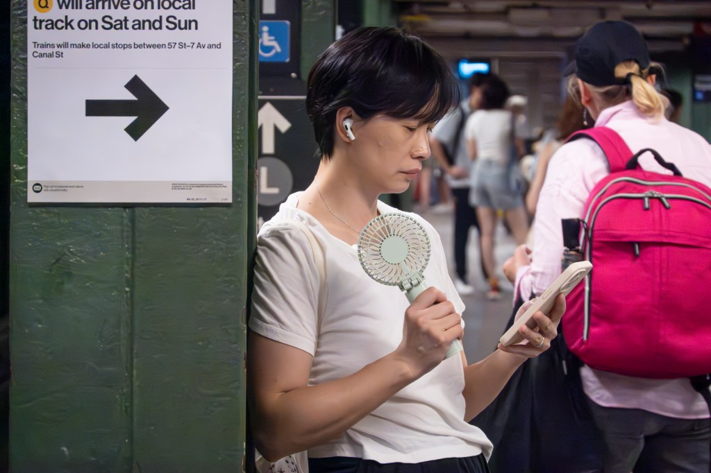 Person using a handheld fan and phone in a subway station.