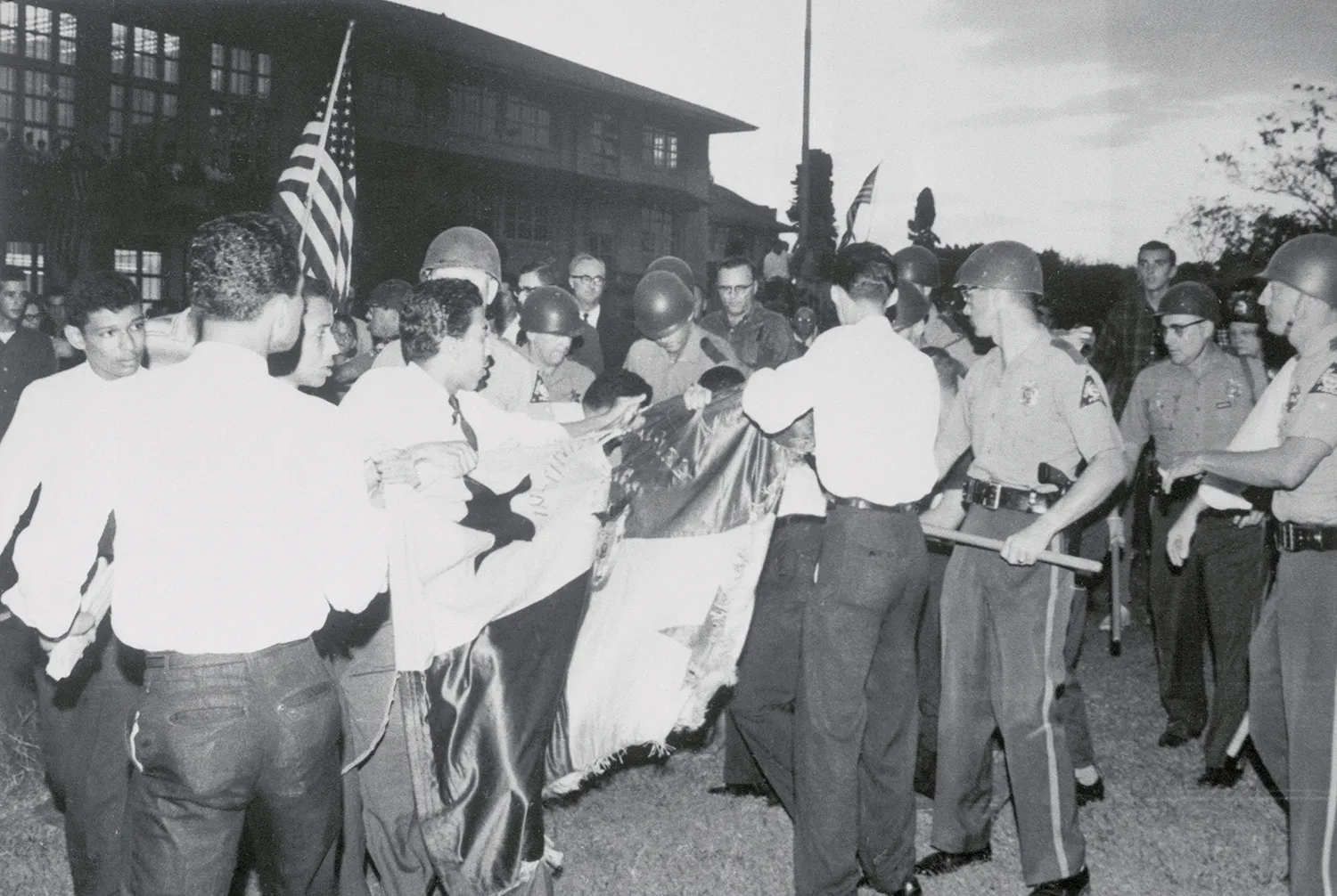 In this black and white photo, dozens of students scuffle in a crowd with police in helmets in a crowd over a flag. A U.S. flag can be seen being waved by someone in the throng at left.