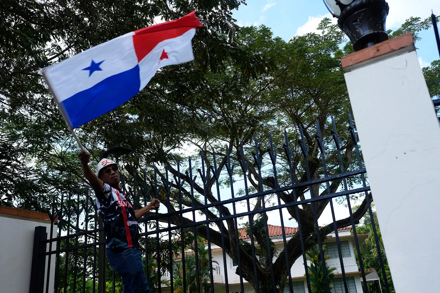 A protester holding a Panama flag scales a fence. A white column is at right, with trees at the roof of a building partly visible behind the fence.