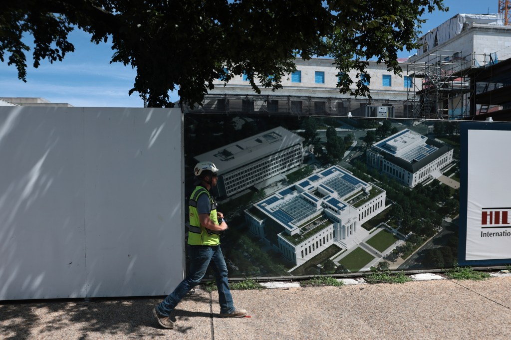 Construction worker walking past a construction site with a rendering of the Federal Reserve building.
