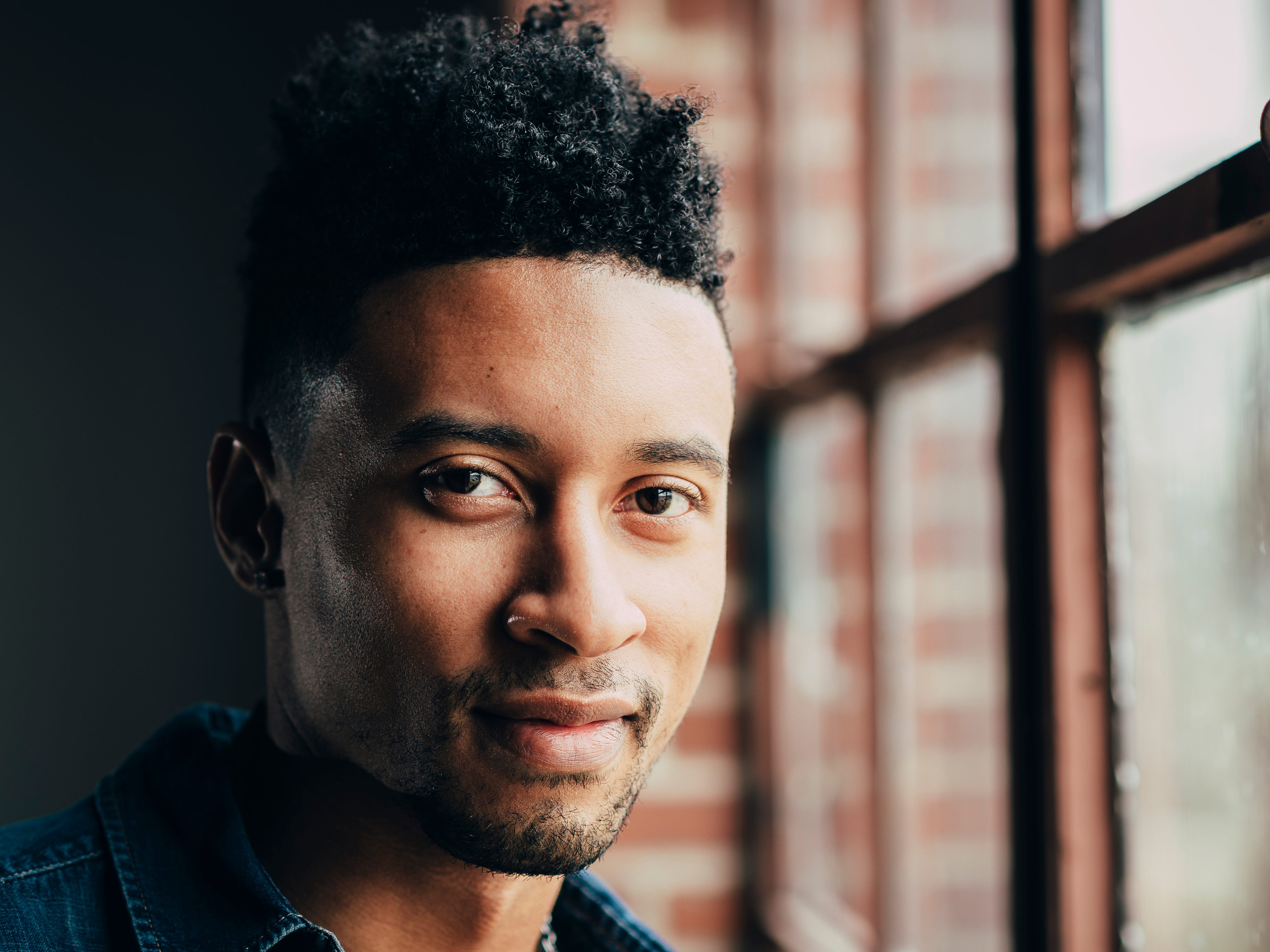An artsy portrait photo of a man looking into the camera next to a window with dramatic lighting
