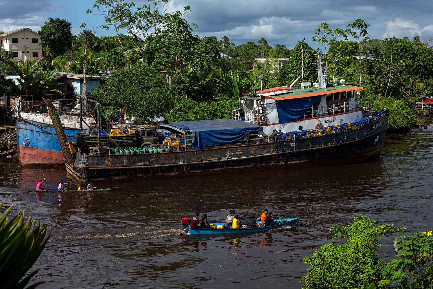 A small motorboat with people in it moves along a river. A largeer boat is seen on the shore. Another small boat is rowed by people down the river.
