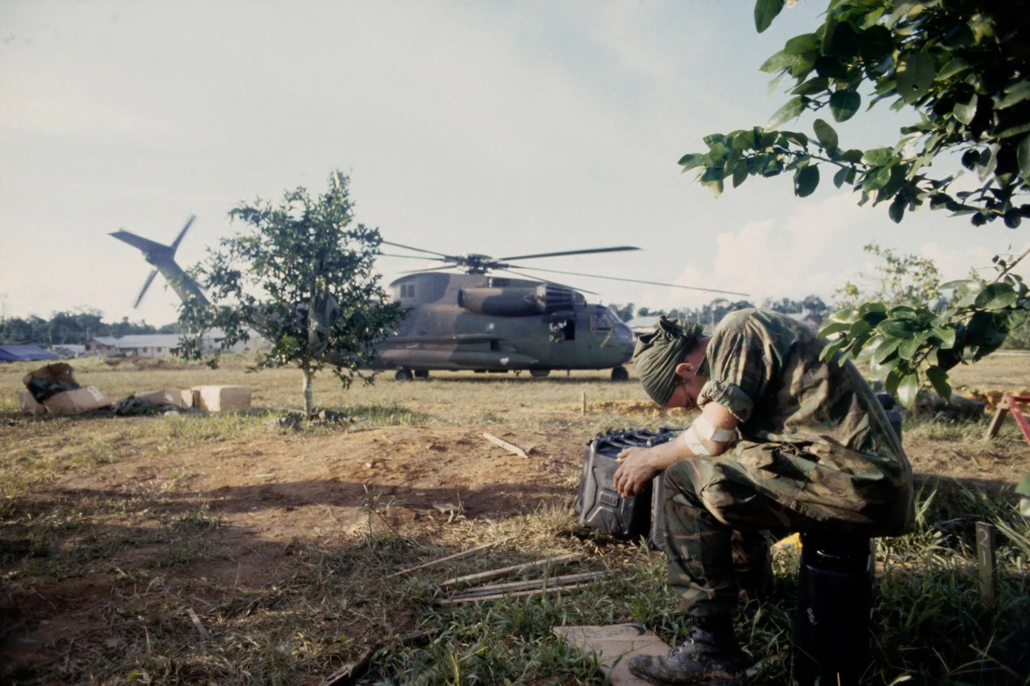 A soldier in camouflage sits with his head bowed. Behind him is a large military helicopter.
