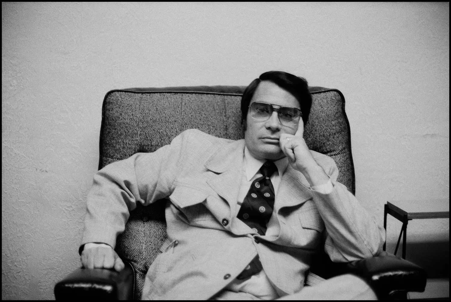 A man in large tinted glasses leans his head on one hand as he sits slouched in a large armchair.