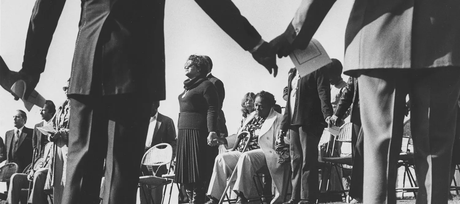 People stand and sit holding hands in a circle in an attitude of prayer and grief.