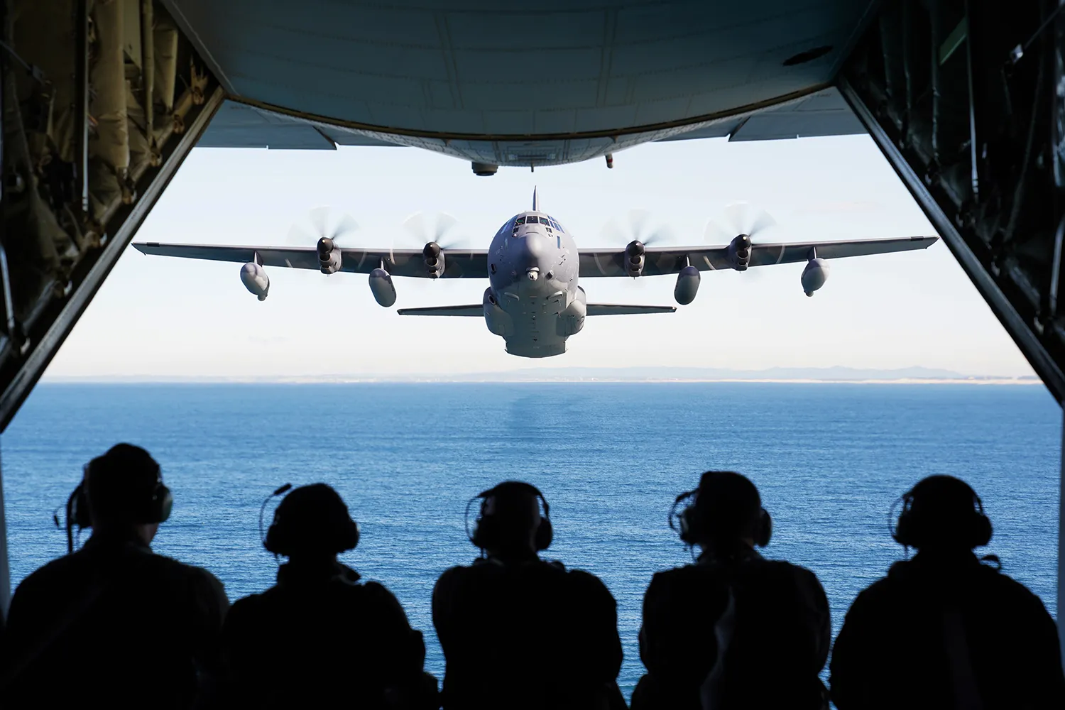 The silhouettes of crew members appear before a blue sea as an aircraft flies ahead toward them.