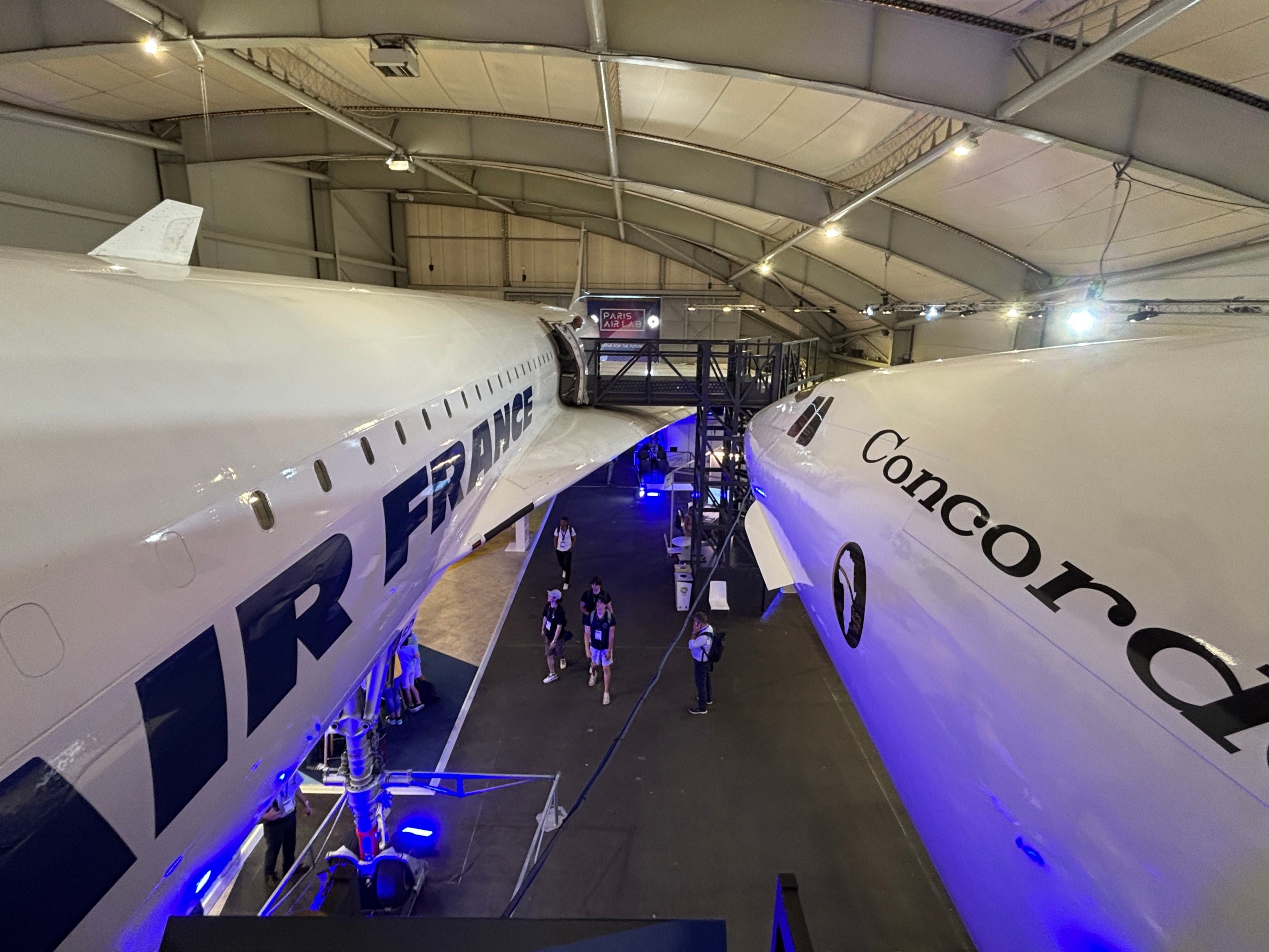 An Air France Concorde Sierra Delta and Concorde 001 side by side above a room of Paris air show attendees