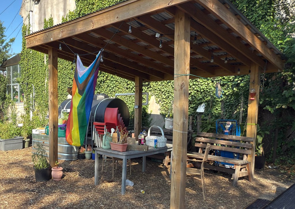 A wooden pergola in a community garden with a pride flag hanging, a table, chairs, and a bench.