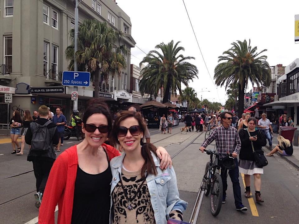 The author and her sister on a city street smiling and wearing sunglasses. There are palm trees, buildings, and people behind them.