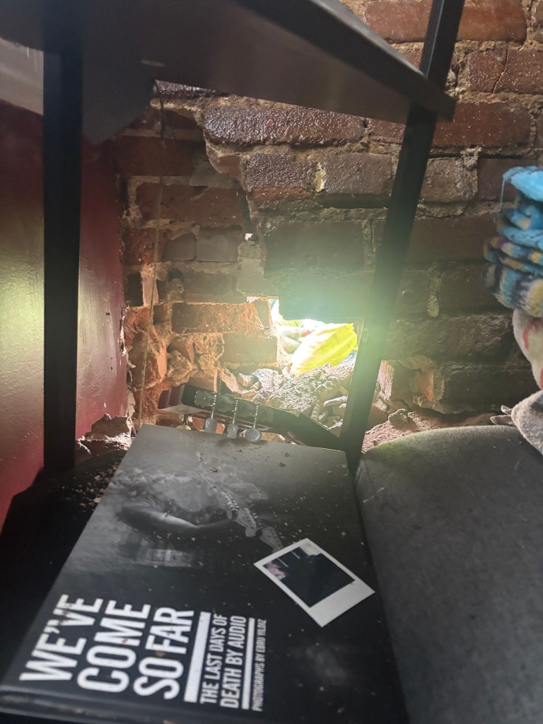 A hole in a brick wall in an apartment, with a guitar and book visible.