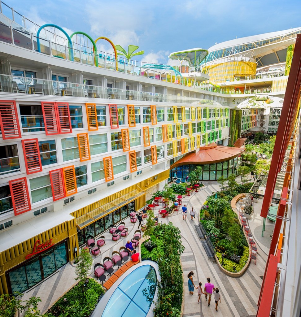 Aerial view of a vibrant cruise ship atrium with colorful buildings, lush greenery, and people walking.