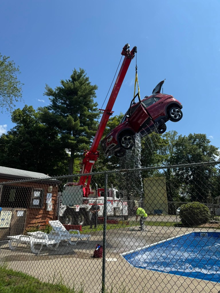 A red car being lifted by a crane over a swimming pool.