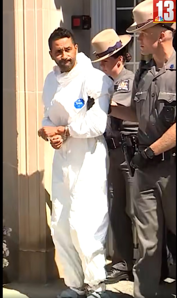 A man in handcuffs and a white protective suit is perp-walked by police.