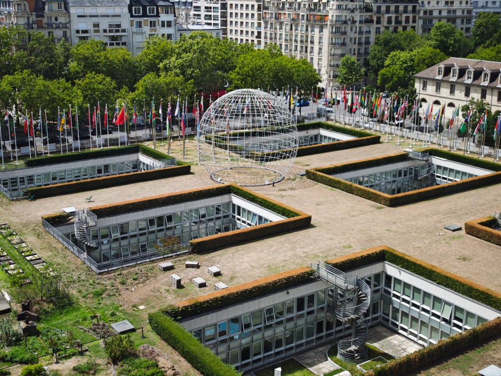 Aerial view of the UNESCO headquarters in Paris, showing the building's architecture, flagpoles with numerous flags, and a large globe structure.