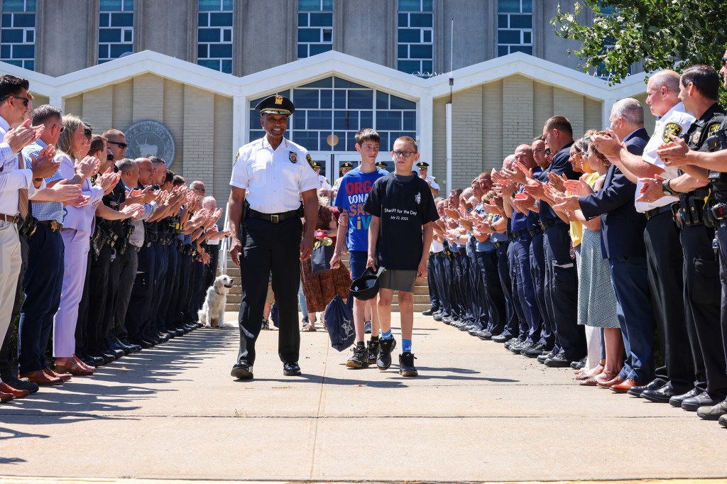 A young boy is sworn in as an honorary sheriff for the day, with a sheriff and a crowd of people applauding.