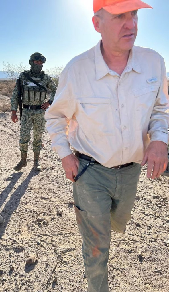 Man in orange hat walking past a soldier in desert terrain.