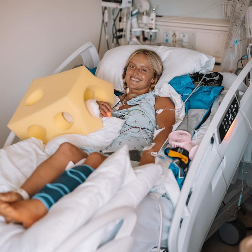 Woman in hospital bed smiling, holding a foam block shaped like a wheel of cheese.