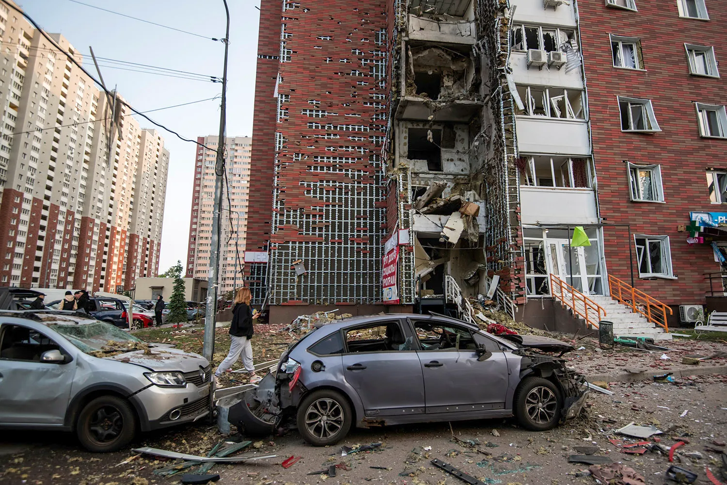 A heavily damaaged apartment building with two damaged cars in the foreground. A person walks by amid debris on the ground.