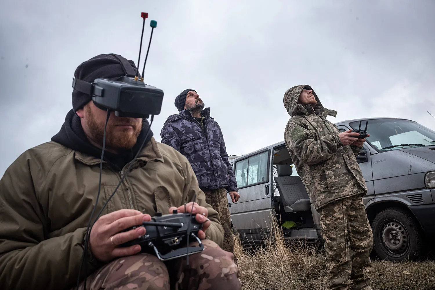 A soldier wears a VR headset and uses a controller. Two men behild him look up at the sky. Behind them is a van with an open door.
