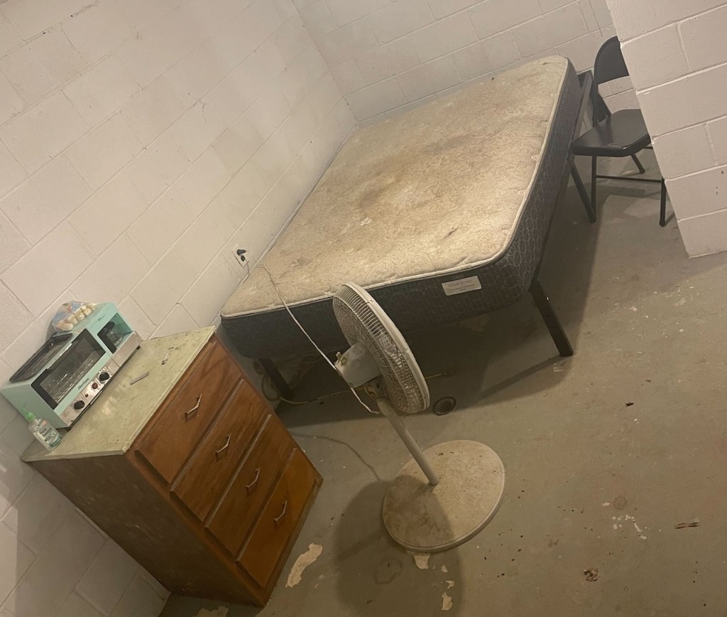Basement room with mattress, fan, dresser, and appliances.