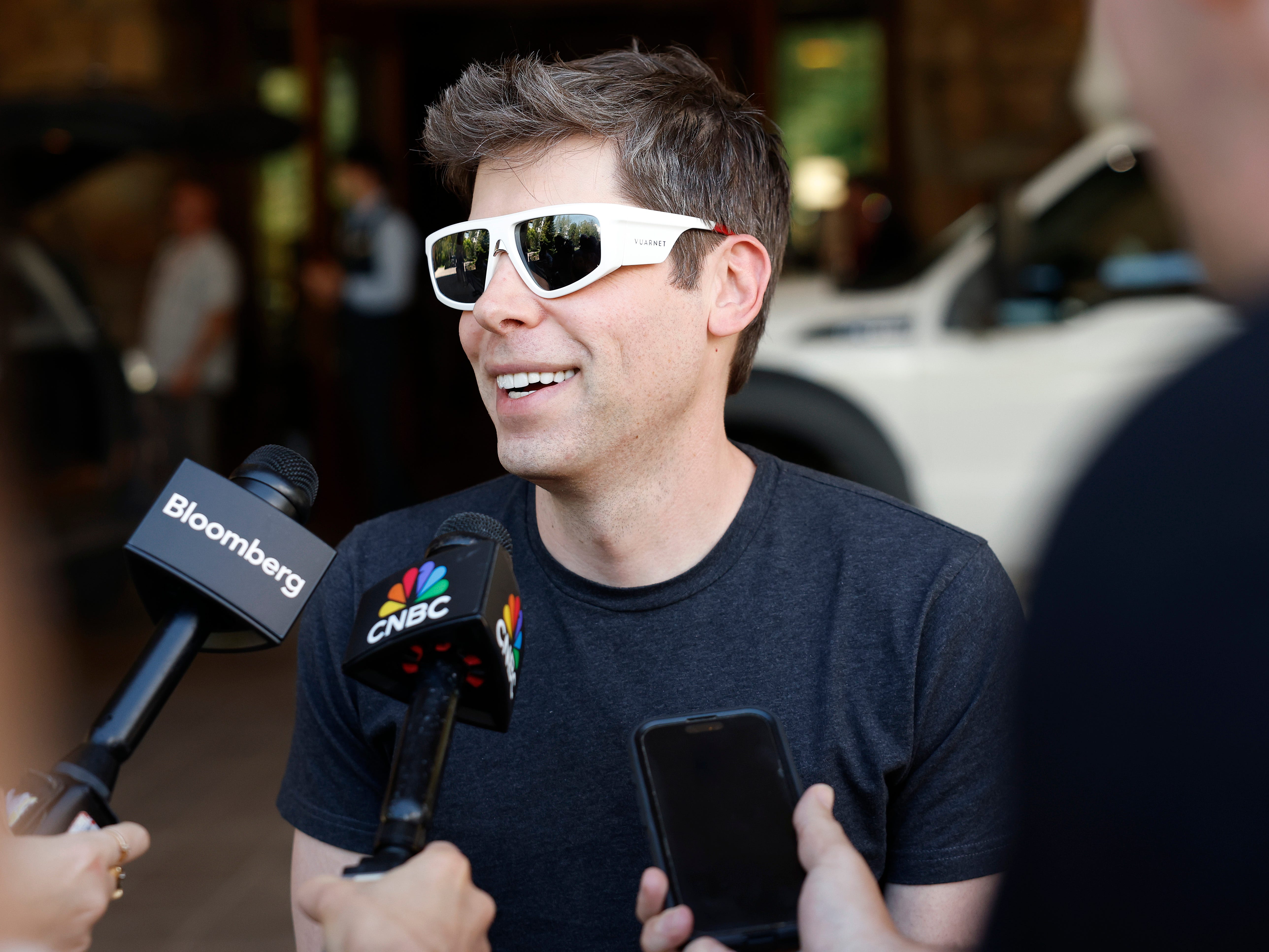 A white man wearing oversize sunglasses speaks to a gaggle of reporters.