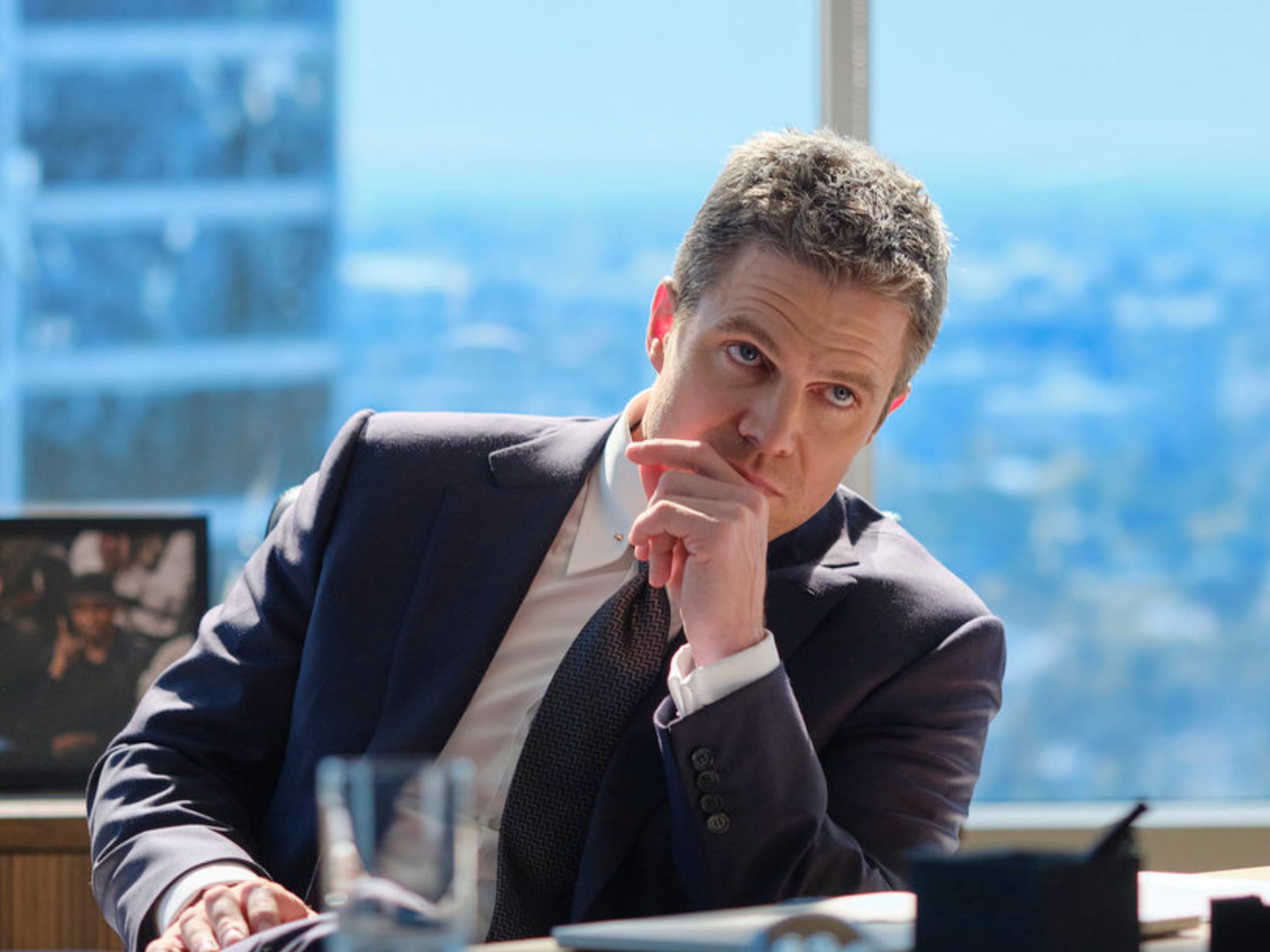 A man with short brown hair is sitting behind a desk in a dark blue suit, white shirt, and patterned tie. Behind him is a large window, and a city can be seen outside. A photo frame on the windowsill is out of focus.