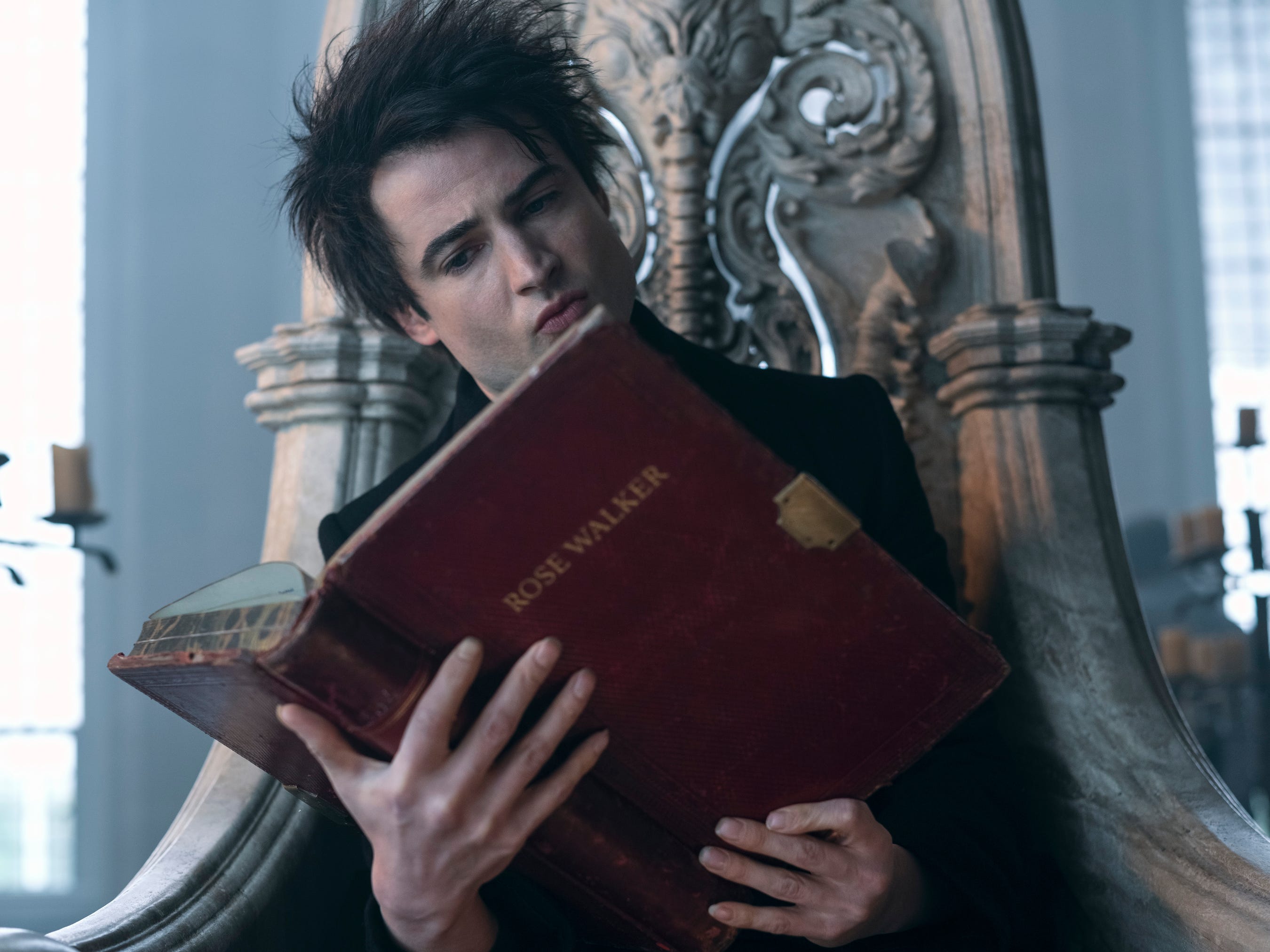 tom sturridge as dream in the sandman, a young man with wild brunette hair, sitting on a stone throne and holding a large, red tome with the text "rose walker" on the front. his hands are splayed across the back of the book and he is reading it intently