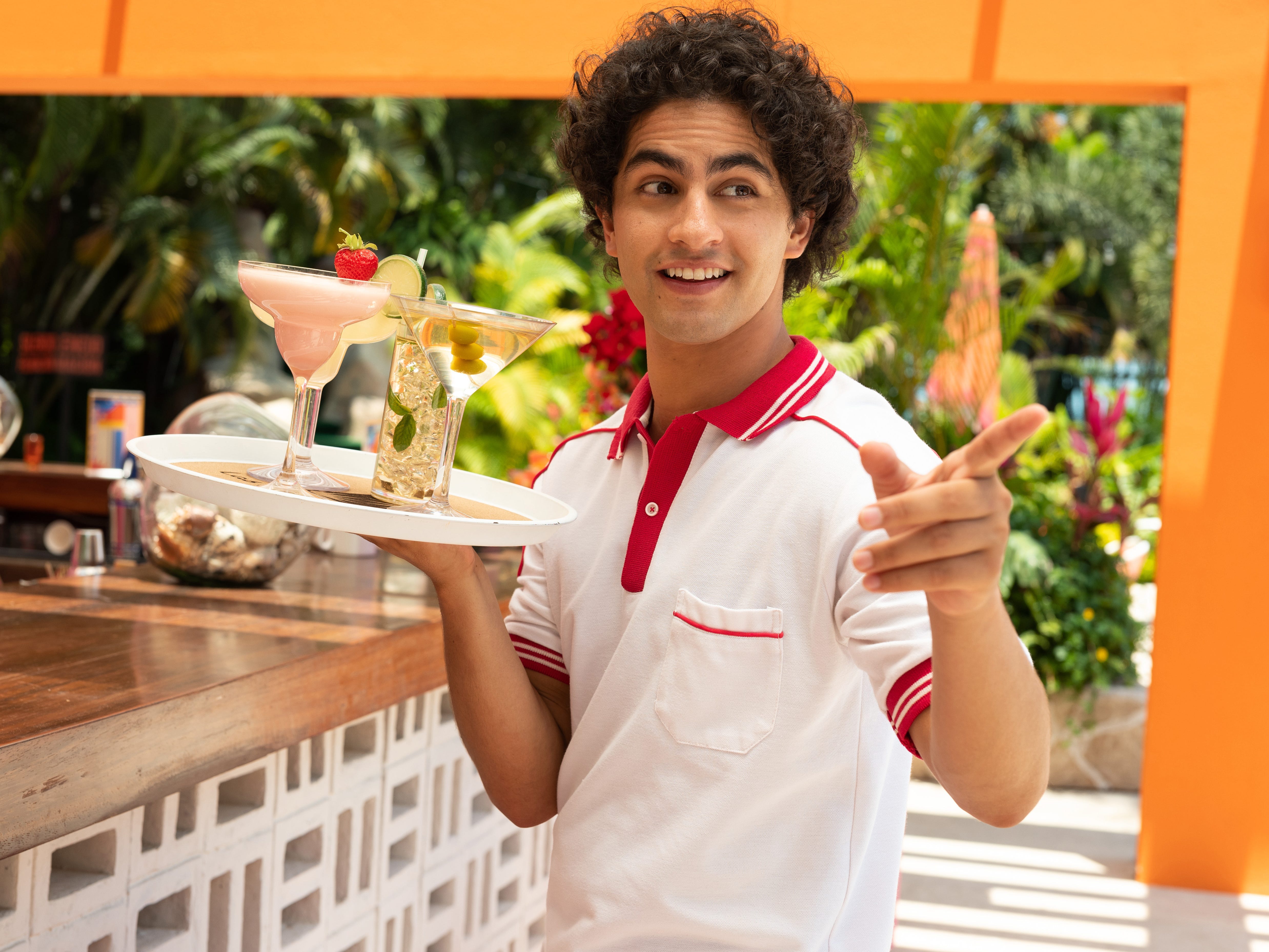 A still of "Acapulco" season one showing a man in a white and red top holding a tray of different alcoholic drinks.