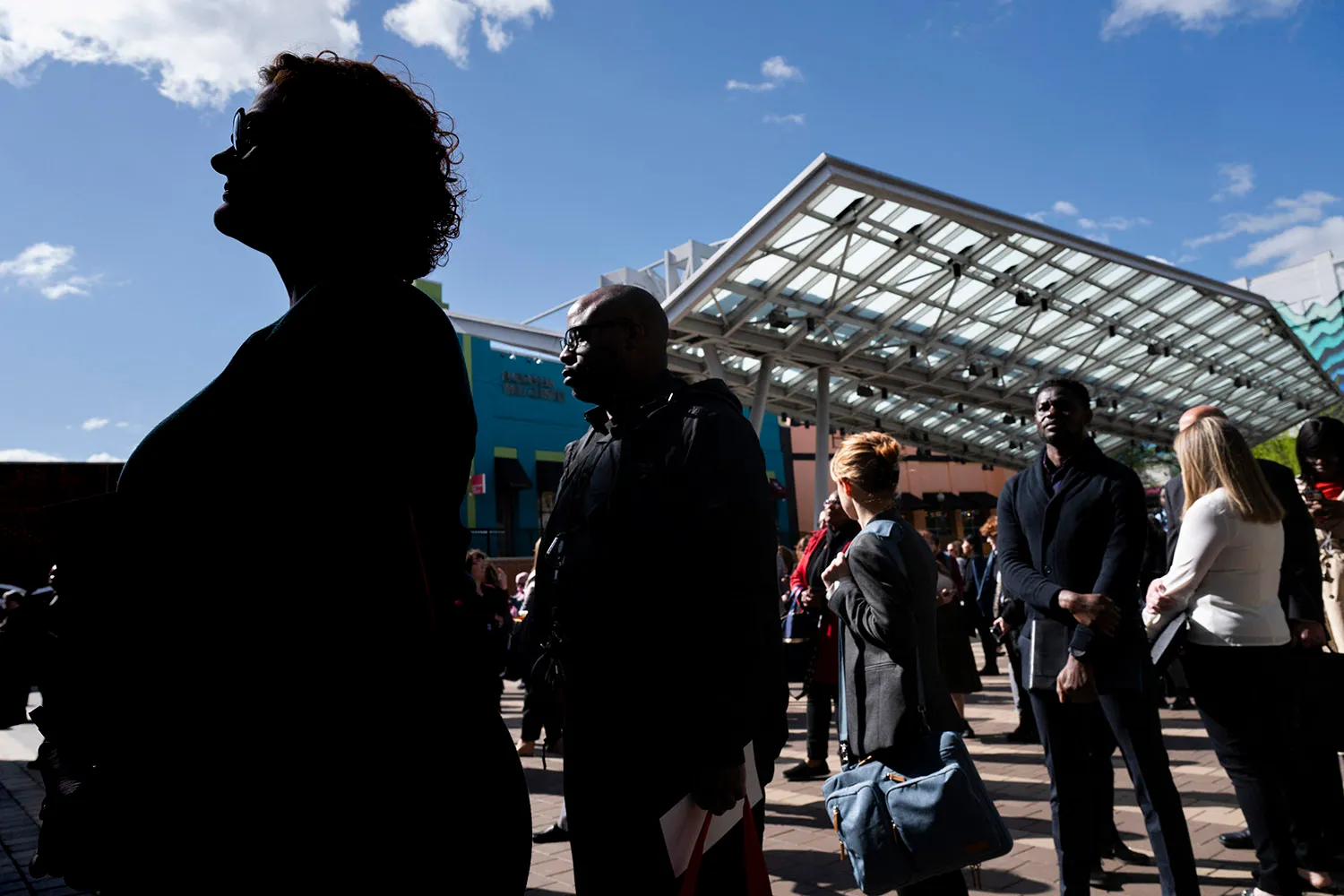 People stand in a line outside near the metal awning of a metro stop beneath a blue sky scattered with a few clouds.