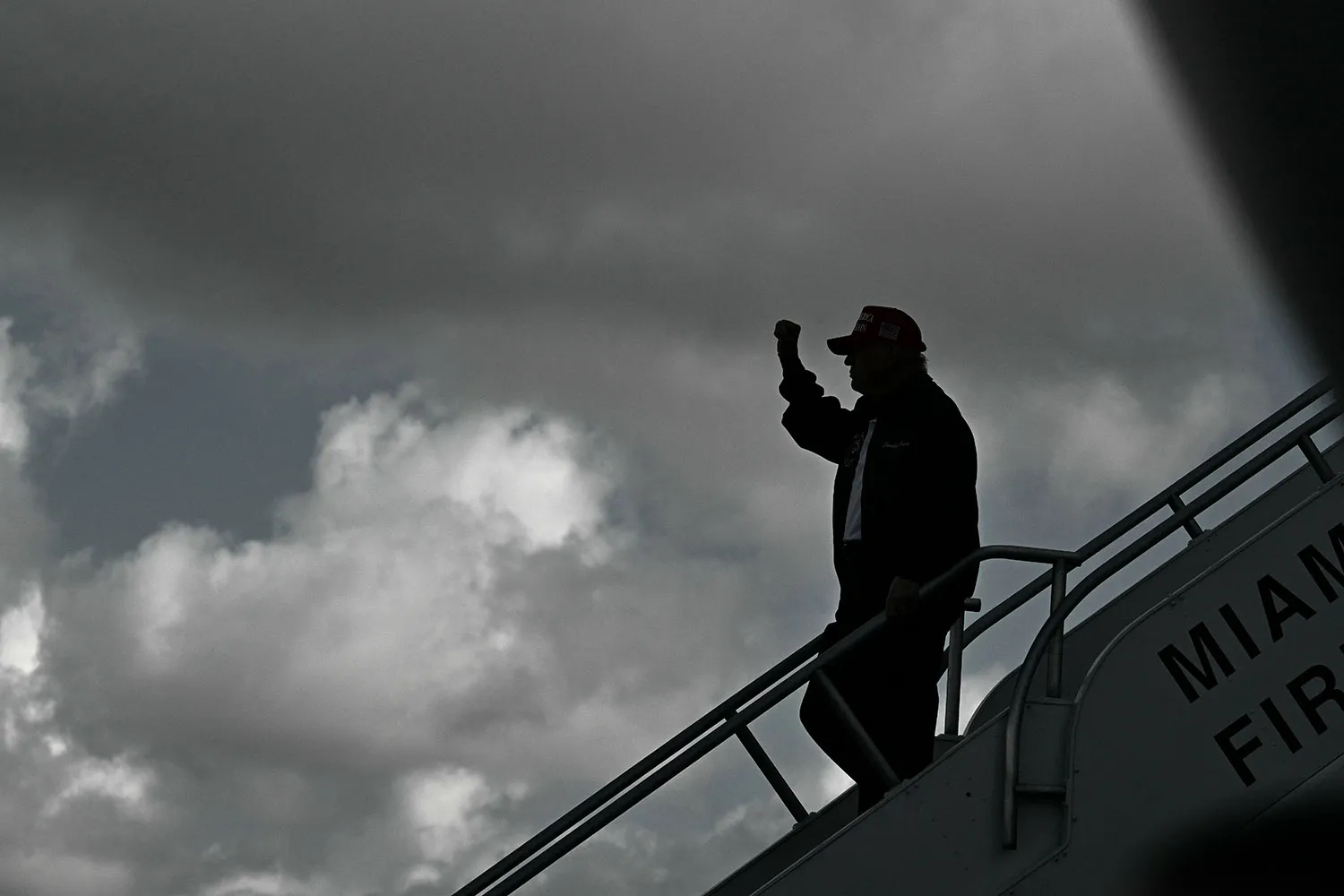 President Trump is seen silhouetted in profile as he raises his fist and walks down the steps of a plane. He stands out sharply against a gray cloudy sky.