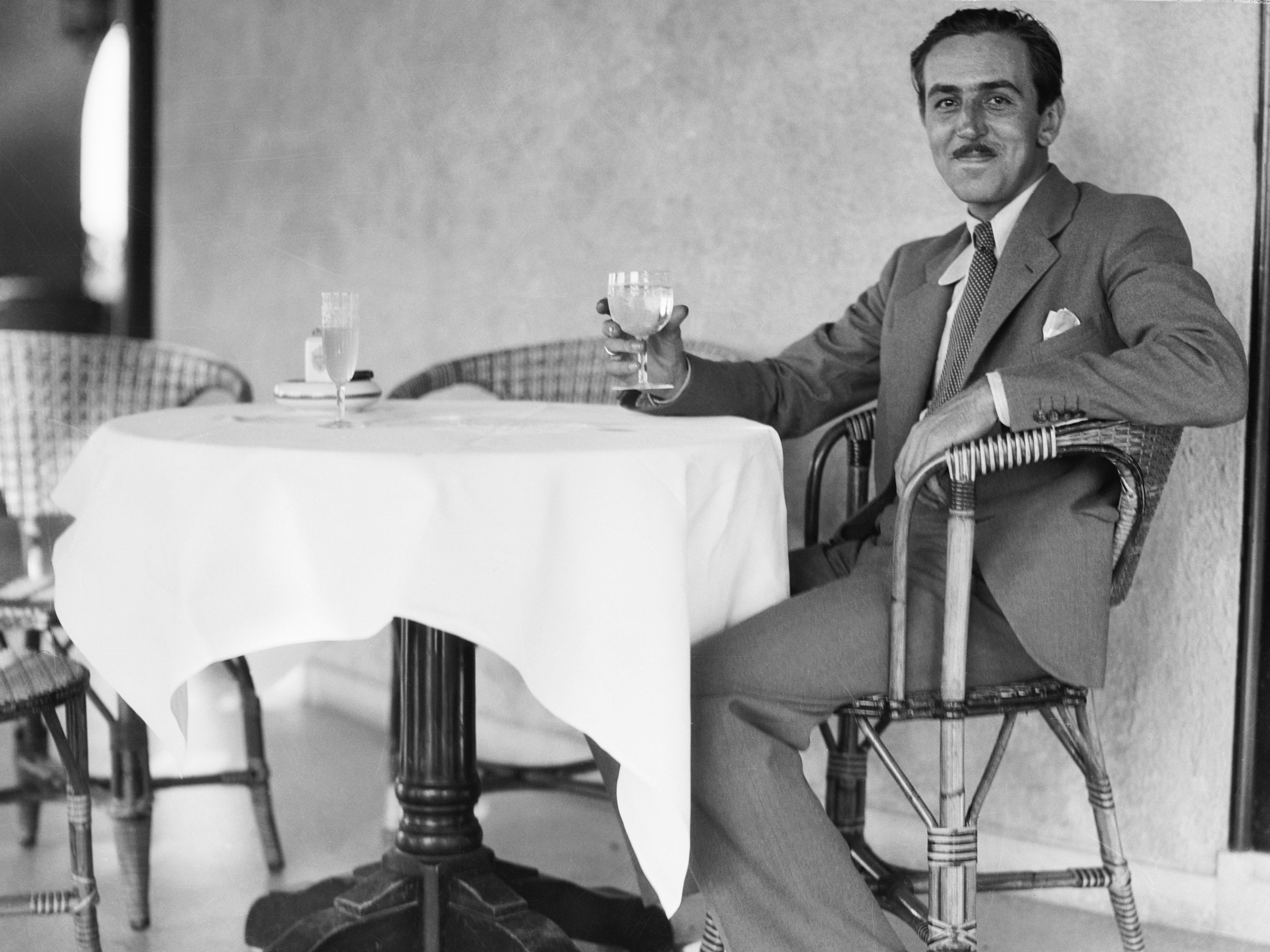 A young Walt Disney sitting in a chair at a table and raising a glass.