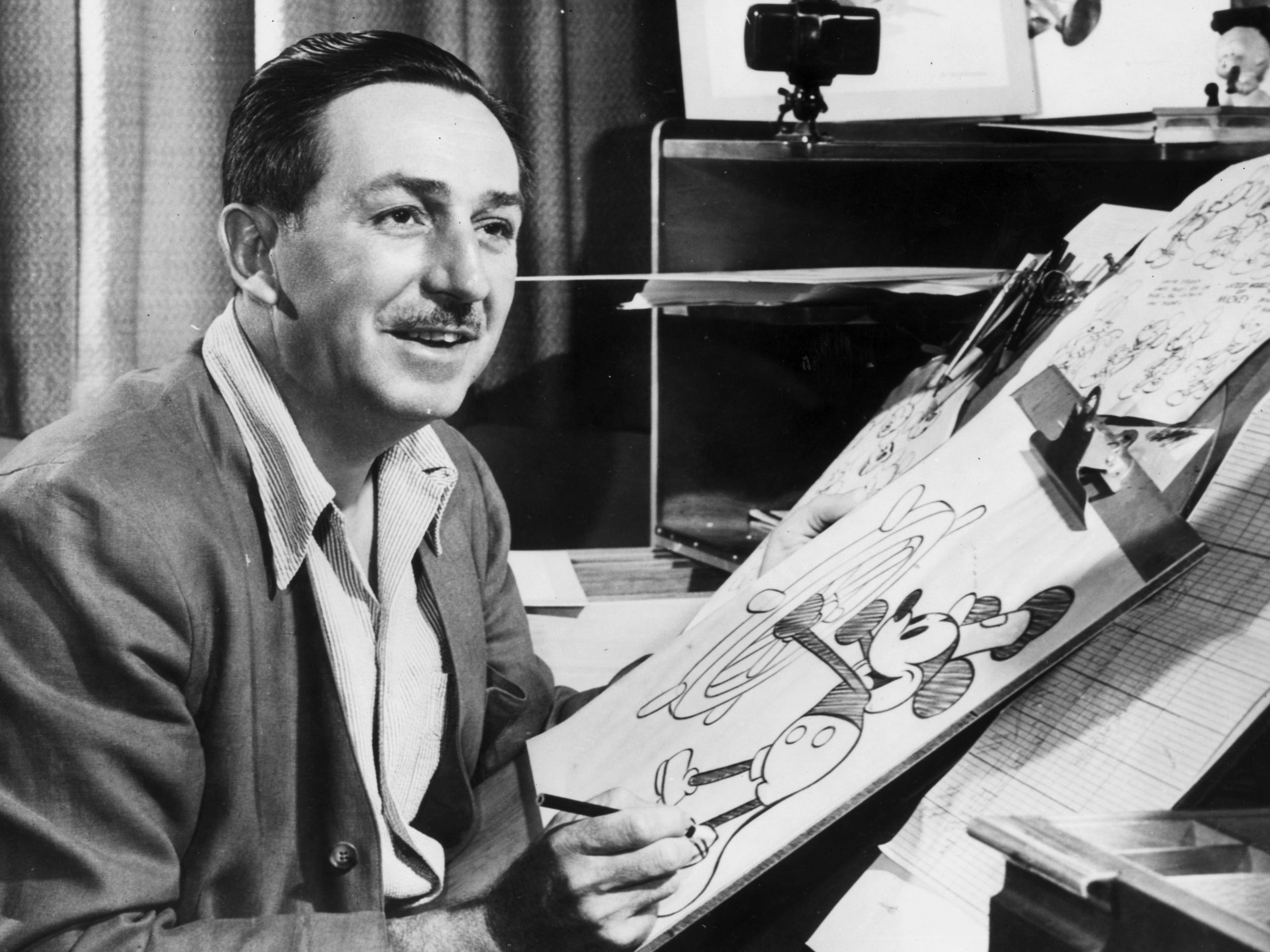 Walt Disney sits at his drawing board in his studio, drawing a sketch of Mickey Mouse.